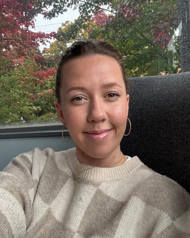 Bre McKinley smiling, wearing a cream sweater, with autumn trees behind her